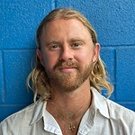 Close-up of a bearded man with long blond hair against a blue wall, wearing a white shirt and gold cross necklace, symbolizing holistic health and personal growth.